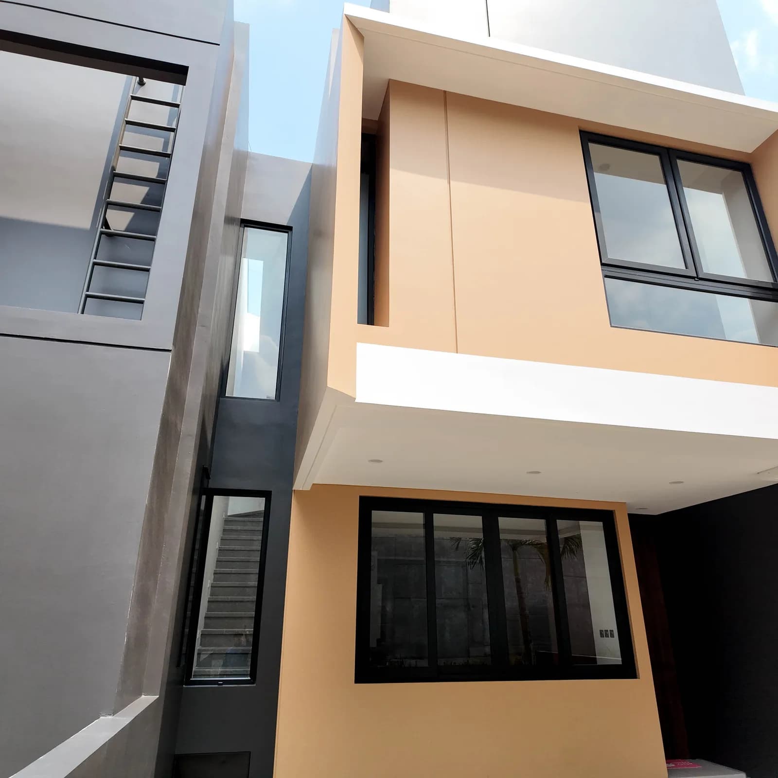 Photograph of a finished house front, tan-beige upper volume and a tall stairwell window under blue sky